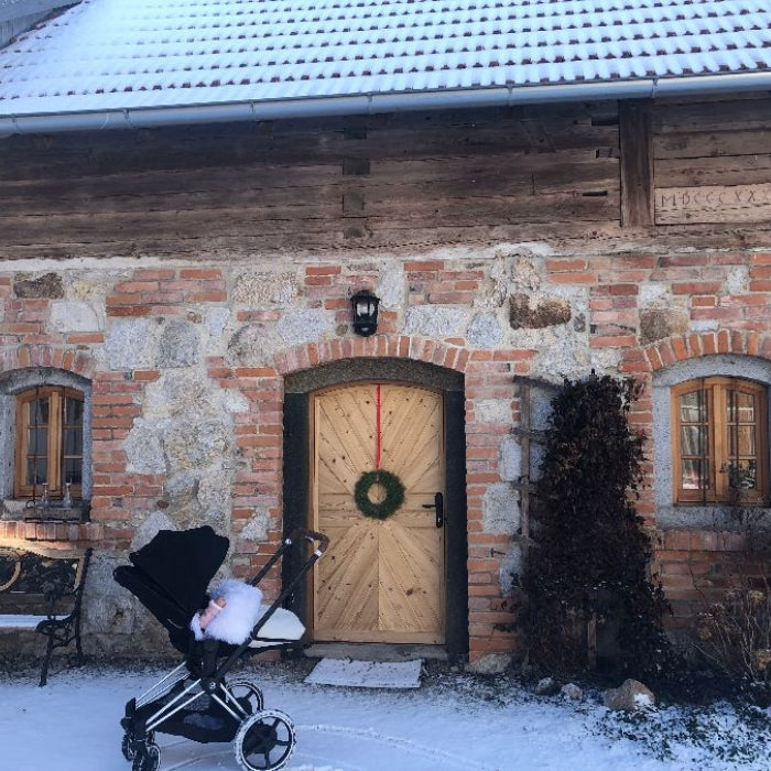 Rustikale Hausfassade der Sabrina Alm Genussmanufaktur im Winter, mit Holztür, weihnachtlichem Kranz und Kinderwagen vor dem Eingang.