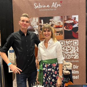 Zwei Personen stehen vor einem Messestand der „Sabrina Alm Genussmanufaktur“ mit großem braunen Banner, QR-Code und Produktfotos im Hintergrund.