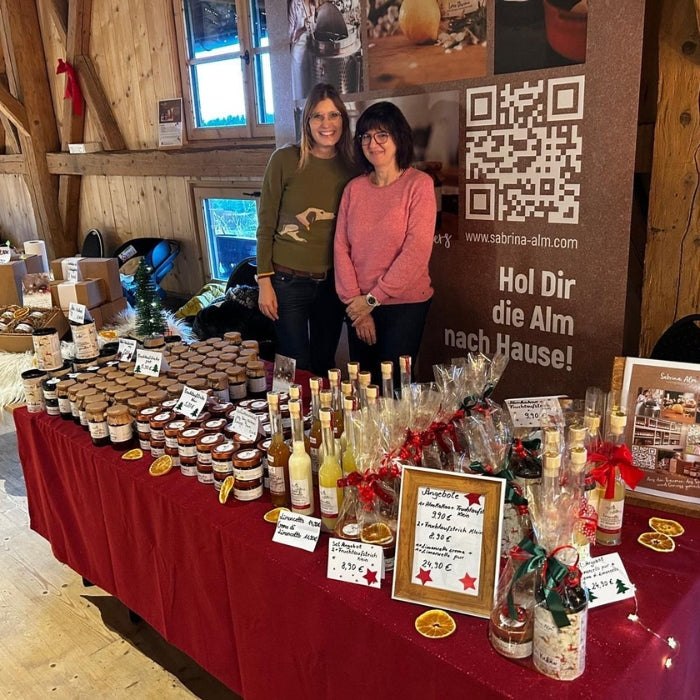 Zwei Frauen stehen hinter einem weihnachtlich dekorierten Stand der „Sabrina Alm Genussmanufaktur“ mit Marmeladen, Likören und verpackten Geschenken auf rotem Tischtuch.