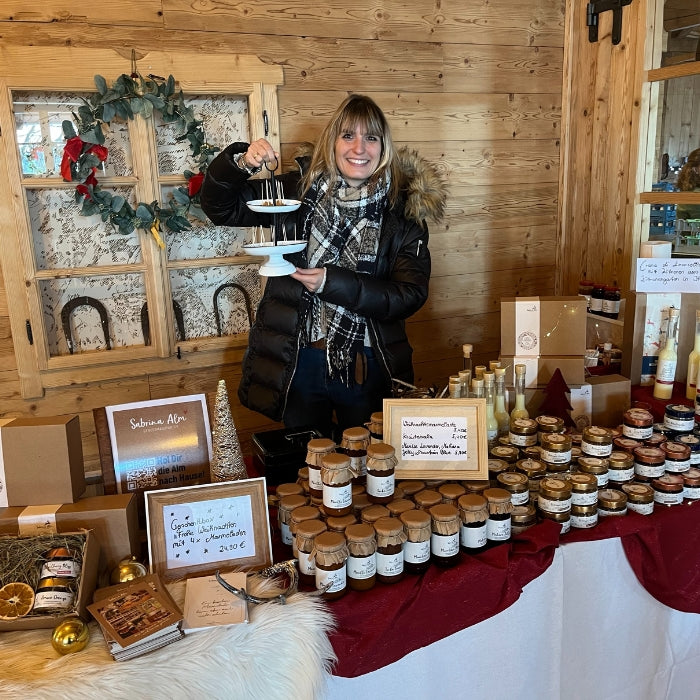 Weihnachtlich dekorierter Marktstand der Sabrina Alm Genussmanufaktur mit handgemachten Marmeladen, Likören und Geschenkboxen – Inhaberin präsentiert Produkte im Holzambiente.